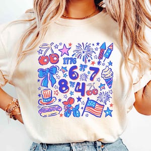Puede incluir: Camiseta color marfil con cuello redondo y dise&ntilde;o gr&aacute;fico colorido. El dise&ntilde;o incluye el n&uacute;mero 1776, fuegos artificiales, estrellas, cerezas, un lazo, un helado, un perrito caliente, un sombrero de copa, un perro globo y una bandera americana.