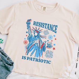 May include: Ivory-colored t-shirt with a graphic of the Statue of Liberty in red and blue, surrounded by stars and fireworks. The text "RESISTANCE IS PATRIOTIC" is printed above and below the image. The shirt is paired with blue jeans.