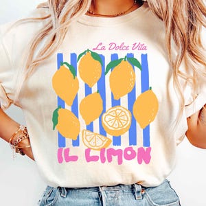 May include: Ivory-colored t-shirt featuring a lemon graphic with the text "La Dolce Vita" and "Il Limon". The design includes yellow lemons, green leaves, and blue vertical stripes. The shirt is a casual wear item.