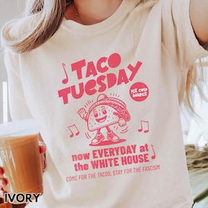 Pode incluir: Camiseta cor marfim com um gr&aacute;fico rosa que diz "TACO TUESDAY" com um personagem de taco de desenho animado. A camisa tamb&eacute;m tem o texto "now EVERYDAY at the WHITE HOUSE" e "COME FOR THE TACOS, STAY FOR THE FASCISM."