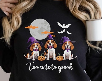 May include: Black sweatshirt featuring three cartoon beagles wearing purple witch hats, sitting in front of a full moon, bats, and pumpkins. The text "Too cute to spook" is written in white script. The word "Black" is in a black oval at the bottom.