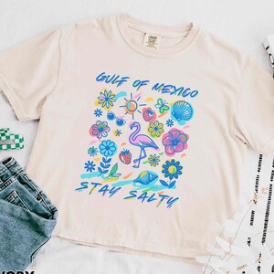 May include: Ivory t-shirt with a graphic print of a flamingo, flowers, and the text "Gulf of Mexico Stay Salty".