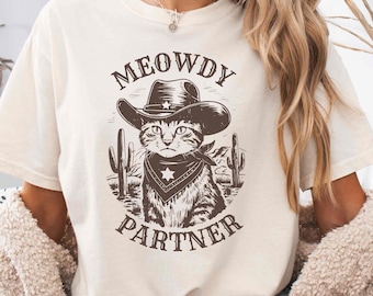 Può includere: Una maglietta bianca con un'illustrazione marrone di un gatto che indossa un cappello da cowboy e un bandana. Il testo "MEOWDY PARTNER" è stampato sulla maglietta.