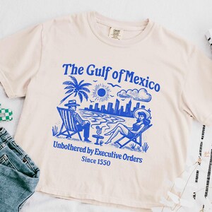 May include: A light beige t-shirt with a blue graphic print of a beach scene with two people sitting on chairs. The text "The Gulf of Mexico" is printed in blue above the graphic. The text "Unbothered by Executive Orders Since 1550" is printed in blue below the graphic.