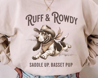 May include: A beige sweatshirt with the text "Ruff & Rowdy" and a cartoon basset hound wearing a cowboy hat and bandana. The text "Saddle Up, Basset Pup" is below the image.