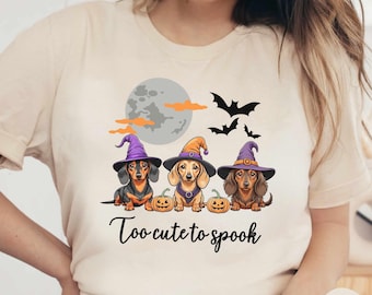 May include: A beige t-shirt with a graphic of three dachshunds wearing witch hats and holding pumpkins. The text "Too cute to spook" is printed below the dachshunds. The t-shirt is on a model wearing blue jeans.