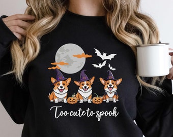 May include: Black sweatshirt with a graphic of three corgis wearing witch hats and holding pumpkins. The text "Too cute to spook" is printed below the corgis.