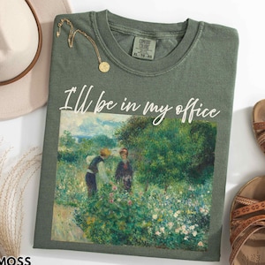 May include: A moss green Comfort Colors t-shirt with the text "I'll be in my office" in white script. The shirt features a painting of two people in a garden. A gold necklace and brown sandals are also visible.
