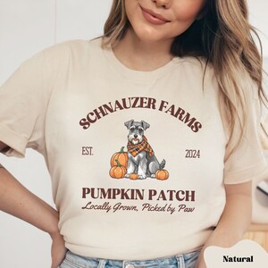 May include: A beige t-shirt with the text "Schnauzer Farms Est. 2024 Pumpkin Patch Locally Grown. Picked by Paw" in brown text. A cartoon miniature schnauzer dog wearing a scarf sits in front of three pumpkins.