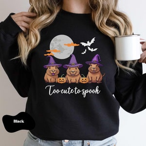 Op de afbeelding: Zwart sweatshirt met een afbeelding van drie cavia's die toverhoeden dragen en pompoenen vasthouden. De tekst "Too cute to spook" staat onder de cavia's.