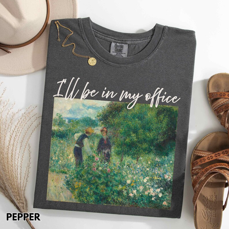 May include: Pepper-coloured Comfort Colors t-shirt with the text "I'll be in my office" above a painting of two people in a garden. A gold necklace and a beige hat are next to the shirt. Brown sandals are on the right.