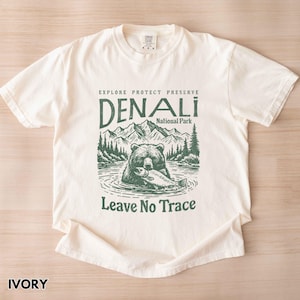 May include: Ivory t-shirt with a green illustration of a bear holding a fish in front of a mountain landscape. The text "Explore Protect Preserve Denali National Park Leave No Trace" is printed on the shirt.
