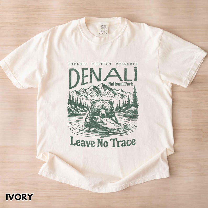 May include: Ivory t-shirt with a green illustration of a bear holding a fish in front of a mountain landscape. The text "Explore Protect Preserve Denali National Park Leave No Trace" is printed on the shirt.