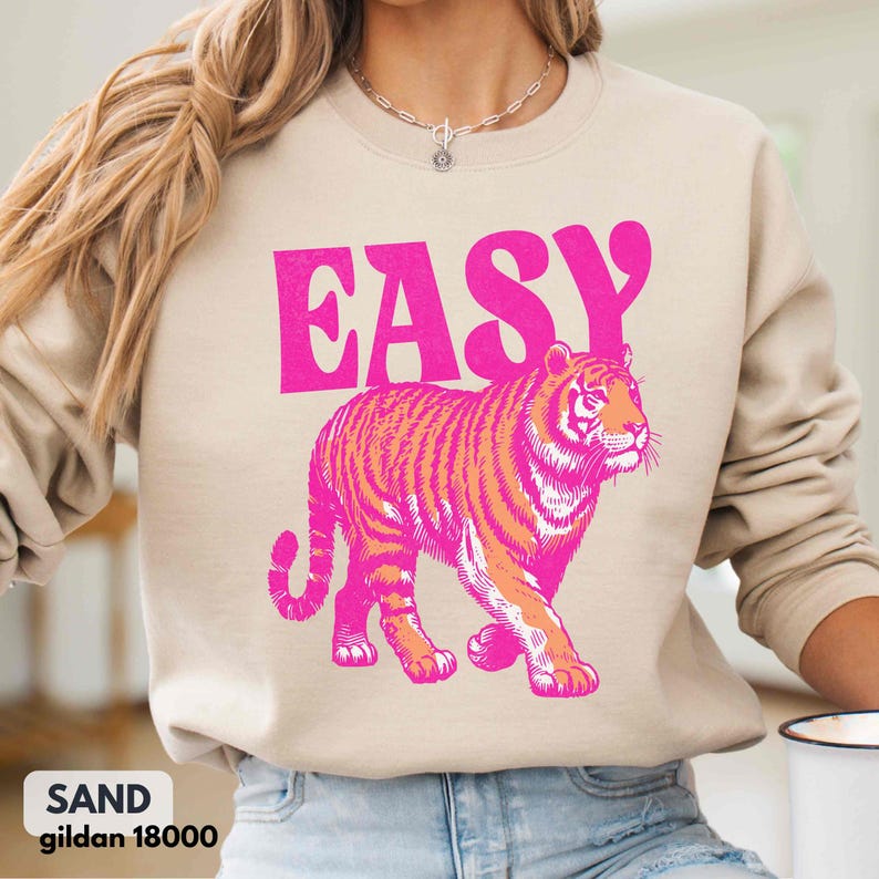 May include: A sand-colored crewneck sweatshirt featuring the word EASY printed in bold hot pink letters above a graphic of a walking tiger. The tiger is illustrated in shades of hot pink and orange, showcasing a detailed striped pattern.