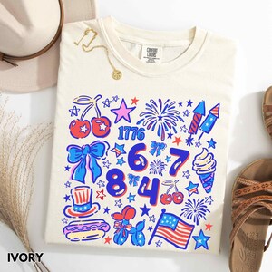 Puede incluir: Camiseta de color marfil con un gr&aacute;fico patri&oacute;tico colorido. El dise&ntilde;o incluye el n&uacute;mero 1776, fuegos artificiales, estrellas, cerezas, lazos, un helado y una bandera estadounidense. La camiseta se presenta con un sombrero, un collar y sandalias.