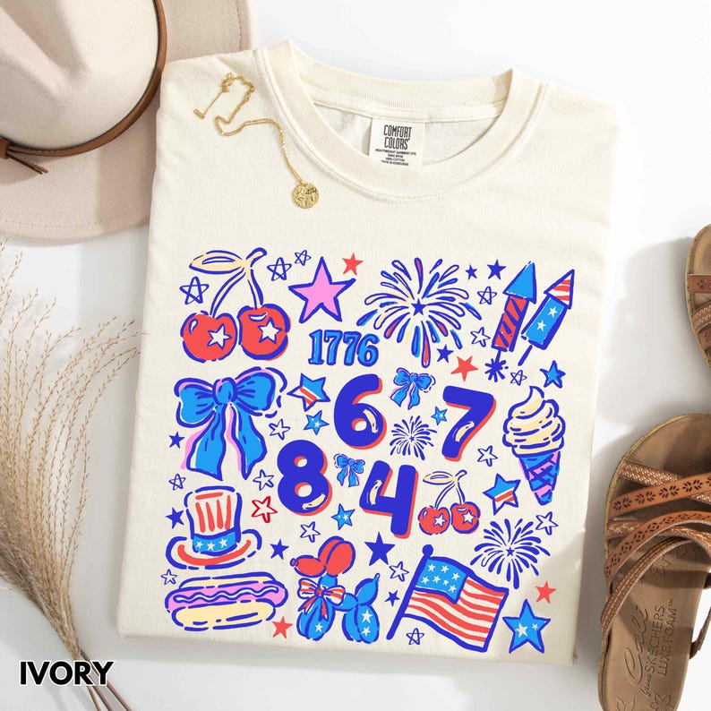 Puede incluir: Camiseta de color marfil con un gr&aacute;fico patri&oacute;tico colorido. El dise&ntilde;o incluye el n&uacute;mero 1776, fuegos artificiales, estrellas, cerezas, lazos, un helado y una bandera estadounidense. La camiseta se presenta con un sombrero, un collar y sandalias.