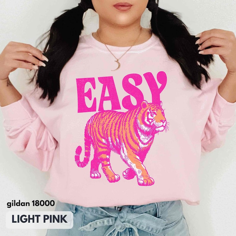 May include: A light pink crewneck sweatshirt featuring the word EASY in bold, bright pink capital letters above a graphic illustration of a tiger in shades of bright pink and orange. The garment has long sleeves and a ribbed neckline.