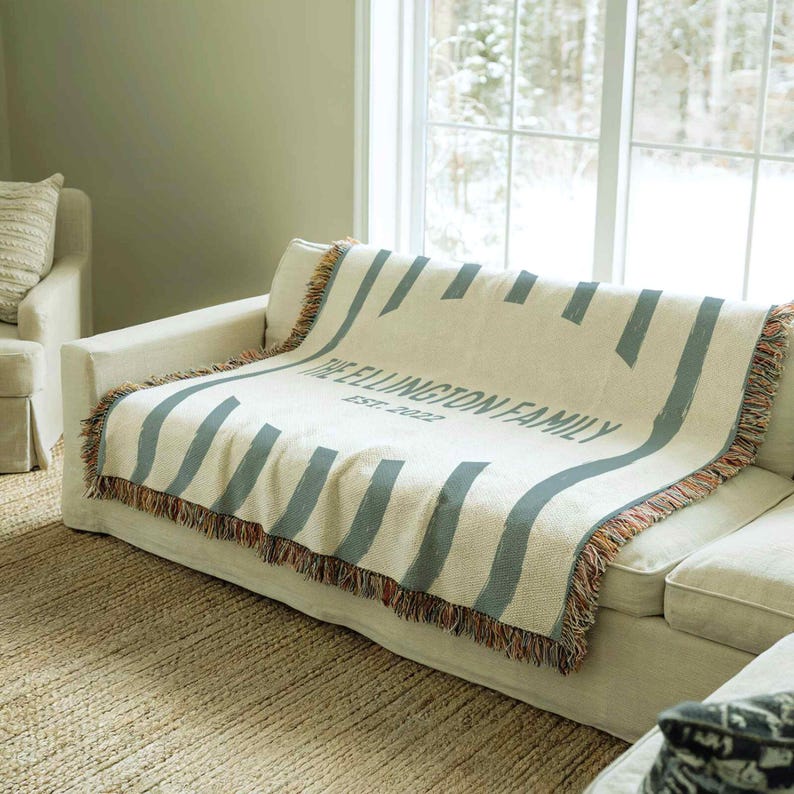 May include: A cream-colored throw blanket with blue-gray stripes and the text "THE ELLINGTON FAMILY EST. 2022" is draped over a white sofa. The blanket has fringed edges with orange and brown hues. The sofa sits on a woven rug.