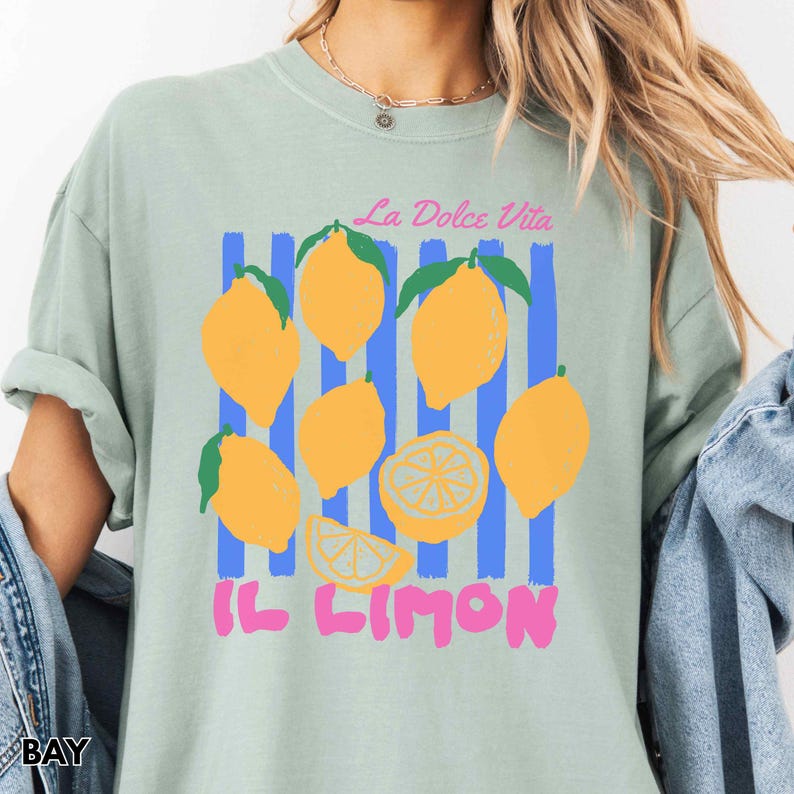 May include: A light green t-shirt featuring a lemon design. The design includes yellow lemons with green leaves, blue vertical stripes, and the text "La Dolce Vita" and "IL LIMON" in pink. The shirt has a relaxed fit.