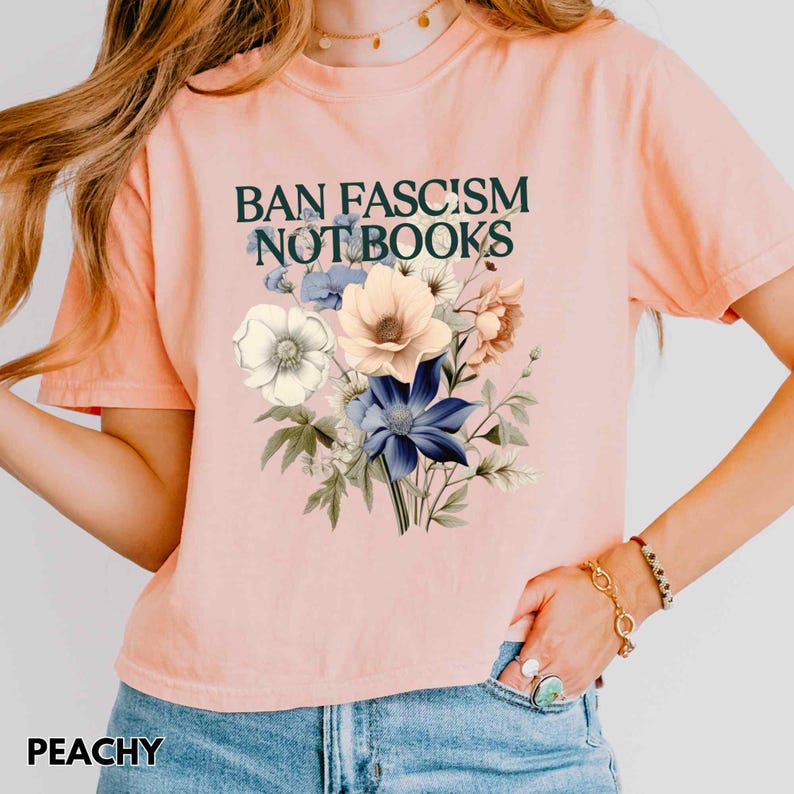 Puede incluir: Camiseta color melocot&oacute;n con el texto "BAN FASCISM NOT BOOKS" sobre un ramo floral. El ramo presenta flores blancas, azules y color melocot&oacute;n con hojas verdes. La palabra "PEACHY" est&aacute; impresa en la parte inferior.
