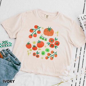Puede incluir: Camiseta de color marfil con un gr&aacute;fico de tomate. El dise&ntilde;o presenta tomates rojos y verdes, junto con formas de estrellas y diamantes amarillos. La camiseta tiene cuello redondo y mangas cortas. La palabra "IVORY" est&aacute; impresa en la parte inferior.