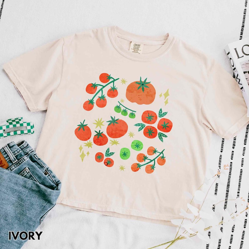 Puede incluir: Camiseta de color marfil con un gr&aacute;fico de tomate. El dise&ntilde;o presenta tomates rojos y verdes, junto con formas de estrellas y diamantes amarillos. La camiseta tiene cuello redondo y mangas cortas. La palabra "IVORY" est&aacute; impresa en la parte inferior.