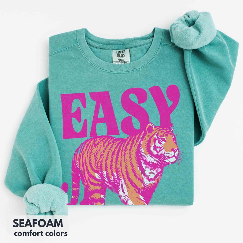 May include: A seafoam green crewneck sweatshirt featuring the word EASY in large, bold, hot pink letters. Below the text is a graphic of a tiger rendered in shades of hot pink and tan. The garment has a Comfort Colors brand label at the neck.