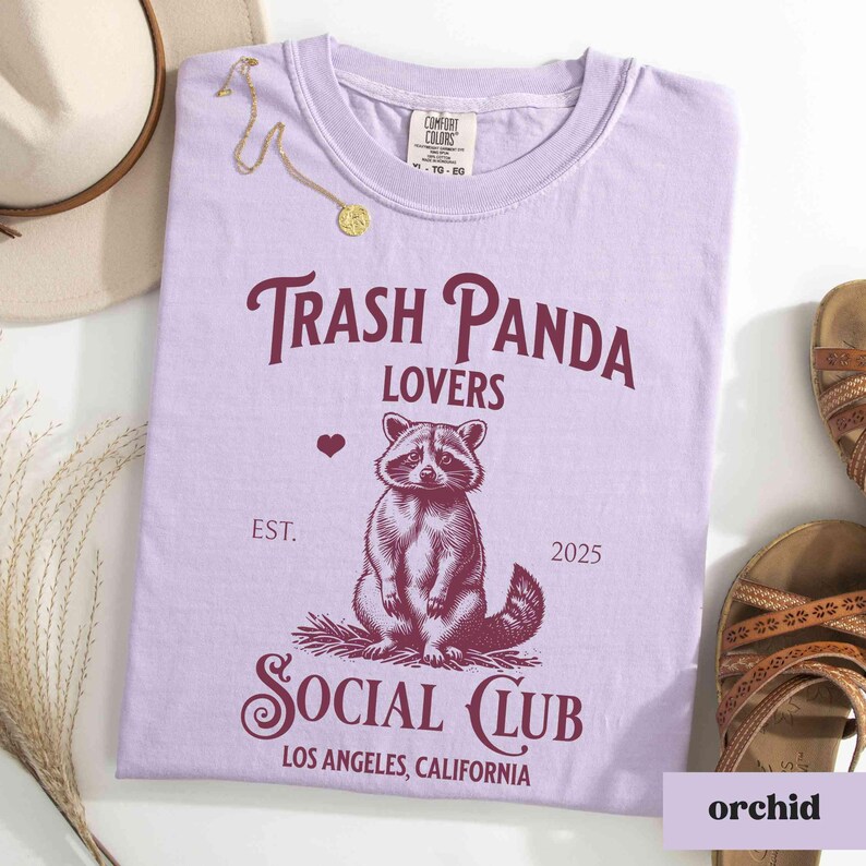 May include: A lavender t-shirt with a graphic of a raccoon and the text "Trash Panda Lovers Social Club Est. 2025 Los Angeles, California".