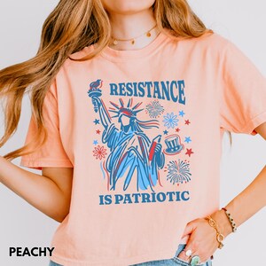 May include: Peach-colored t-shirt featuring a graphic of the Statue of Liberty in blue and red, with the words "RESISTANCE IS PATRIOTIC." The design includes fireworks, stars, and a top hat. The word "PEACHY" is at the bottom left.
