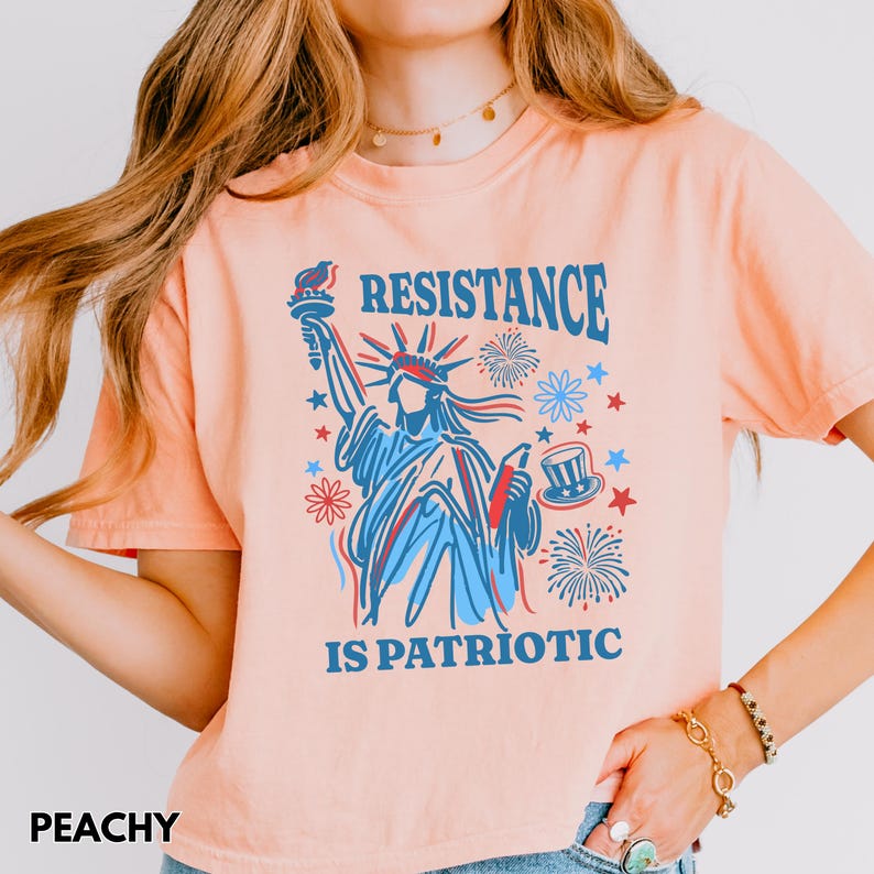 May include: Peach-colored t-shirt featuring a graphic of the Statue of Liberty in blue and red, with the words "RESISTANCE IS PATRIOTIC." The design includes fireworks, stars, and a top hat. The word "PEACHY" is at the bottom left.