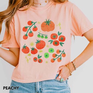 Puede incluir: Camiseta color melocot&oacute;n con un dise&ntilde;o de tomate colorido. El gr&aacute;fico incluye tomates rojos y verdes de varios tama&ntilde;os, as&iacute; como ramas de tomates con hojas verdes. La palabra "PEACHY" est&aacute; impresa en la parte inferior de la camiseta.