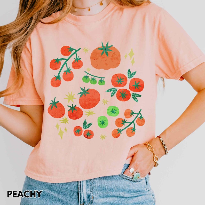 Puede incluir: Camiseta color melocot&oacute;n con un dise&ntilde;o de tomate colorido. El gr&aacute;fico incluye tomates rojos y verdes de varios tama&ntilde;os, as&iacute; como ramas de tomates con hojas verdes. La palabra "PEACHY" est&aacute; impresa en la parte inferior de la camiseta.