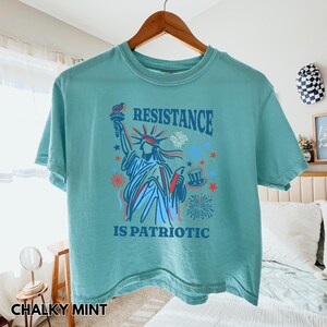 May include: A chalky mint green t-shirt with a graphic of the Statue of Liberty holding a torch, with the words "RESISTANCE IS PATRIOTIC" in blue and red. The shirt is hanging on a wooden hanger.