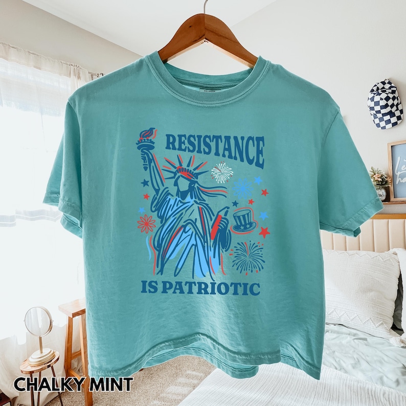 May include: A chalky mint green t-shirt with a graphic of the Statue of Liberty holding a torch, with the words "RESISTANCE IS PATRIOTIC" in blue and red. The shirt is hanging on a wooden hanger.