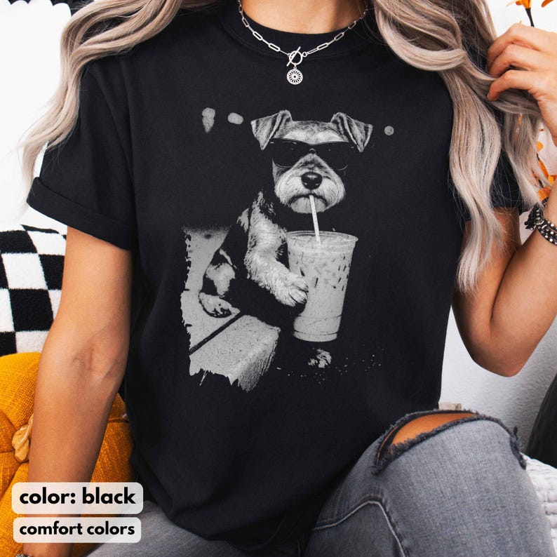 Pode incluir: Camiseta preta com um gr&aacute;fico em tons de cinza de um c&atilde;o Schnauzer usando &oacute;culos de sol e bebendo de um copo transparente de caf&eacute; gelado. As palavras "color: black" e "comfort colors" s&atilde;o impressas na parte inferior.