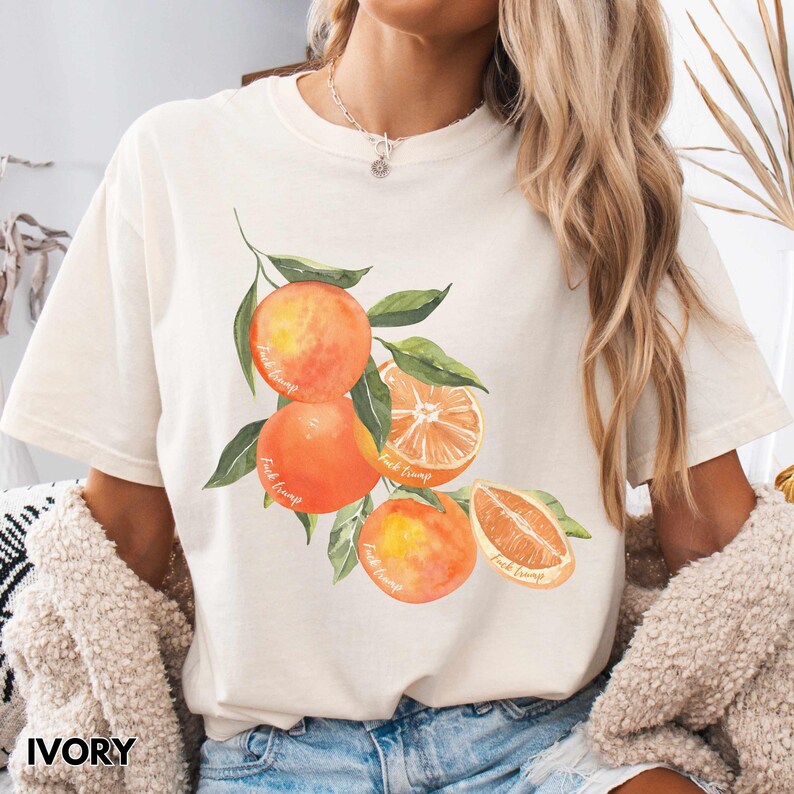 以下が含まれることがあります： オレンジが枝に描かれた水彩画のイラストがプリントされた白いTシャツ。オレンジは鮮やかなオレンジ色で、緑の葉が描かれています。各オレンジには「Fruit Loops」という文字が書かれています。
