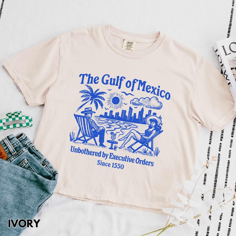 May include: A light beige t-shirt with a blue graphic print of a beach scene with two people sitting on chairs. The text "The Gulf of Mexico" is printed in blue above the graphic. The text "Unbothered by Executive Orders Since 1550" is printed in blue below the graphic.