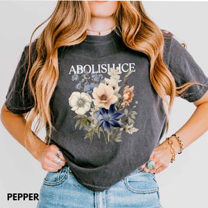 May include: Dark gray t-shirt with the text "ABOLISH ICE" above a floral bouquet. The bouquet features white, peach, blue, and yellow flowers. The word "PEPPER" is printed at the bottom of the shirt.