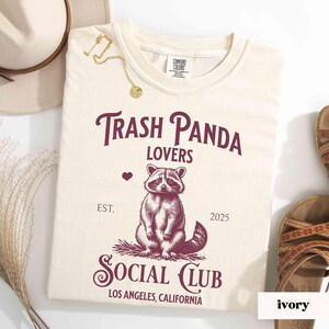 May include: A cream colored t-shirt with a burgundy graphic of a raccoon with the text "Trash Panda Lovers" and "Social Club" with the text "Est. 2025" and "Los Angeles, California".