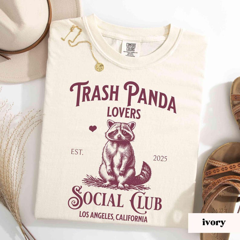 May include: A cream colored t-shirt with a burgundy graphic of a raccoon with the text "Trash Panda Lovers" and "Social Club" with the text "Est. 2025" and "Los Angeles, California".