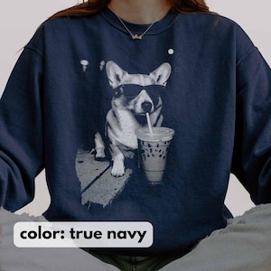 May include: True navy blue sweatshirt featuring a black and white graphic of a corgi wearing sunglasses and drinking from a clear iced coffee cup with a straw. The sweatshirt has a crew neck and long sleeves. The text "color: true navy" is at the bottom.