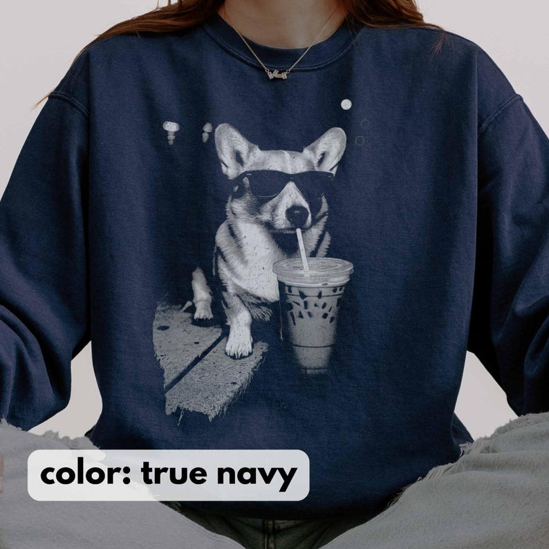 May include: True navy blue sweatshirt featuring a black and white graphic of a corgi wearing sunglasses and drinking from a clear iced coffee cup with a straw. The sweatshirt has a crew neck and long sleeves. The text "color: true navy" is at the bottom.