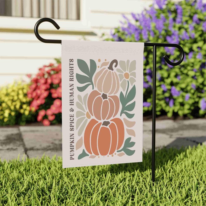 May include: A cream-colored garden flag with a stacked pumpkin design in shades of orange and brown, accented with green leaves and white flowers. The flag reads "PUMPKIN SPICE & HUMAN RIGHTS" in a vertical font. The flag is displayed on a black metal stand in a garden setting.