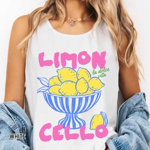 May include: White tank top with a colorful graphic design. The design features the words "LIMON CELLO" in pink, with a bowl of yellow lemons and the words "la dolce vita" in green. The word "WHITE" is printed in white.