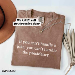 May include: Espresso-colored t-shirt with the text "If you can't handle a joke, you can't handle the presidency." The shirt also has the text "We ONLY sell progressive gear." A silver necklace with a pendant is on the shirt.