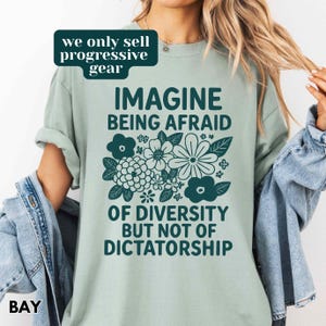 May include: A light green t-shirt with the text "IMAGINE BEING AFRAID OF DIVERSITY BUT NOT OF DICTATORSHIP" in dark green. The shirt features a floral design. The words "we only sell progressive gear" are in a green box.