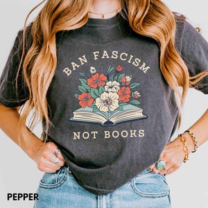 Op de afbeelding: Donkergrijs t-shirt met de tekst "BAN FASCISM NOT BOOKS" boven een illustratie van een open boek met bloemen. De bloemen zijn rood, wit en groen. Het shirt heeft de kleur peper.