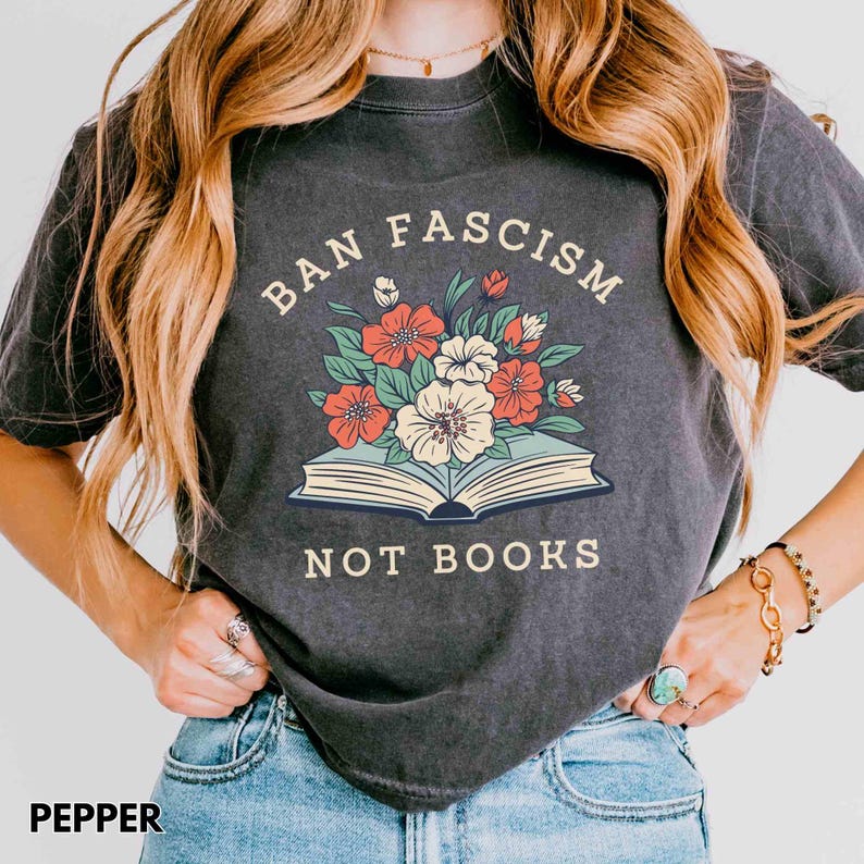 Op de afbeelding: Donkergrijs t-shirt met de tekst "BAN FASCISM NOT BOOKS" boven een illustratie van een open boek met bloemen. De bloemen zijn rood, wit en groen. Het shirt heeft de kleur peper.