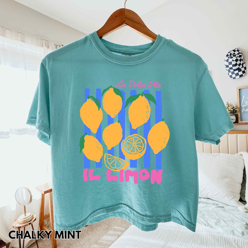 May include: A teal short-sleeve t-shirt with a lemon graphic. The graphic includes yellow lemons, blue stripes, and the text "La Dolce Vita" and "Il Limon" in pink. The shirt is hanging on a wooden hanger.