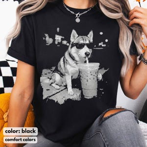 May include: Black t-shirt featuring a grayscale graphic of a husky wearing sunglasses and drinking from a clear iced coffee cup with a straw. The words "color: black" and "comfort colors" are printed at the bottom of the shirt.